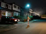 A man stands in the middle of a street at night facing a red car with its lights on.