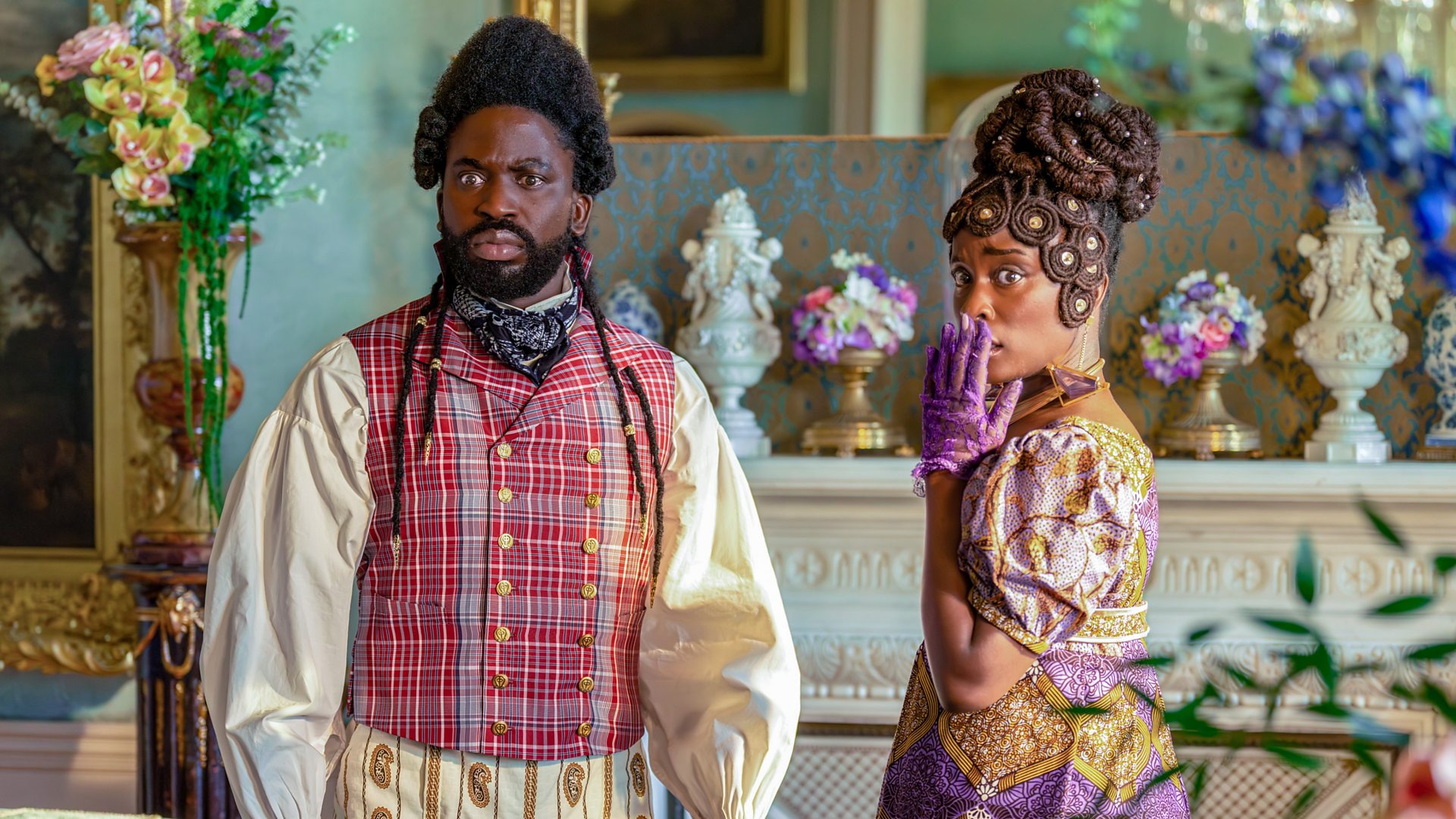 A black man and woman stand in brightly colored, regency era clothing. The woman has her hand covering her mouth.