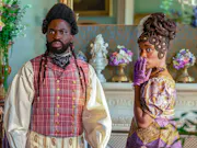 A black man and woman stand in brightly colored, regency era clothing. The woman has her hand covering her mouth.