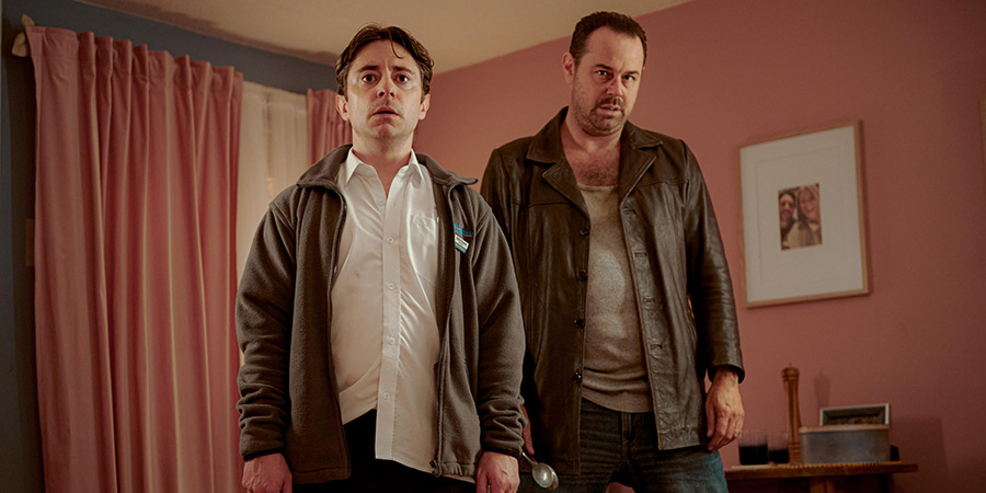 Two white men stand in a pink room wearing brown jackets.