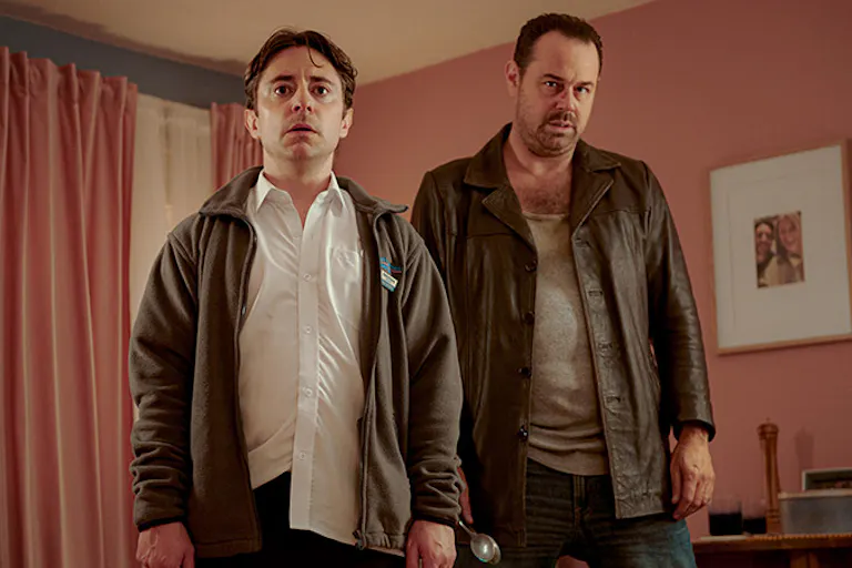 Two white men stand in a pink room wearing brown jackets.