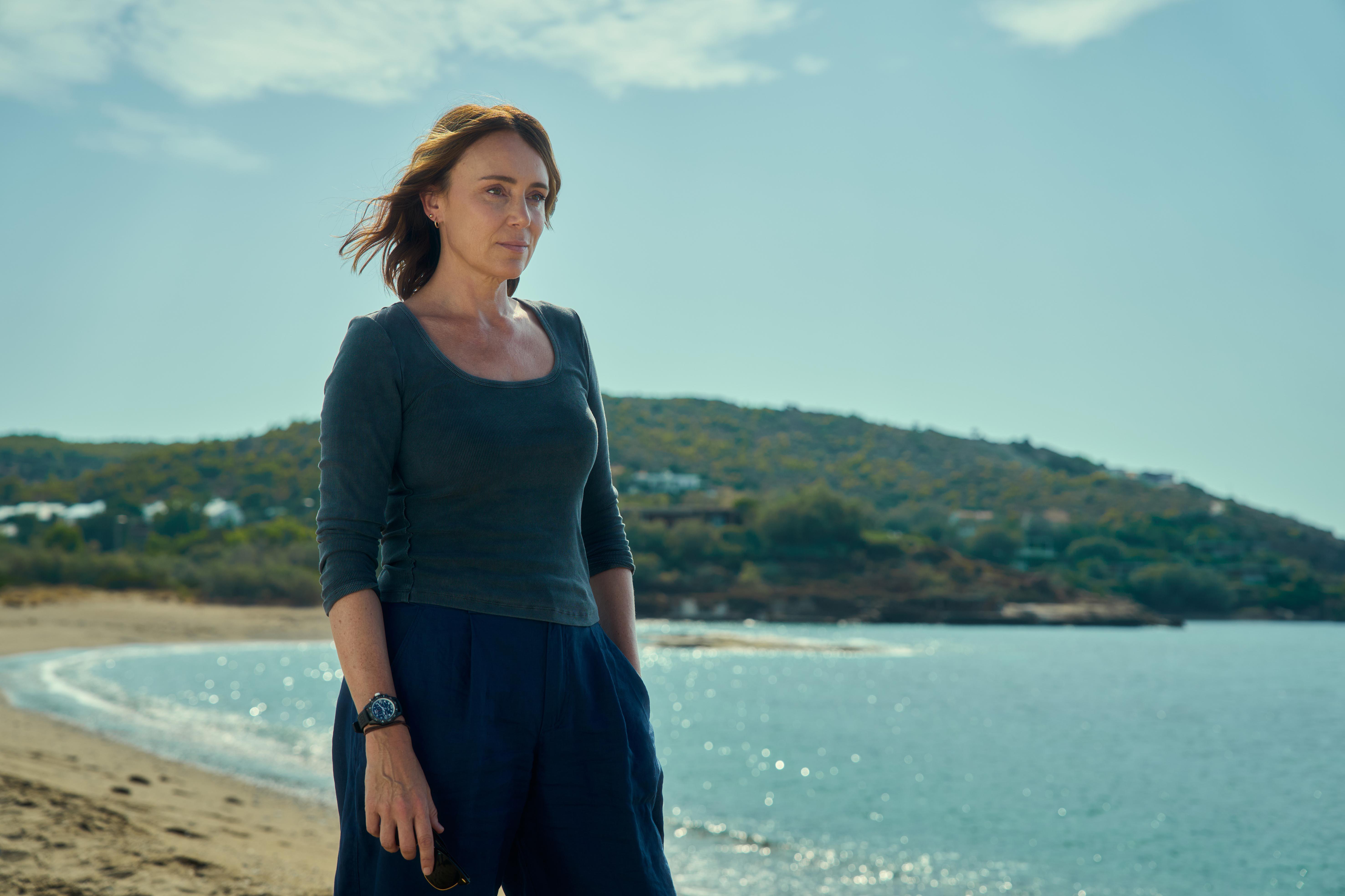 A white woman stands on a beach in a teal top and dark pants.