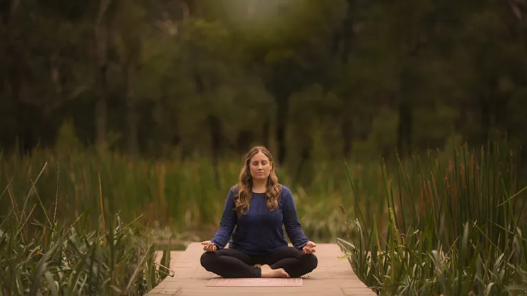 A white woman sitting crossed legged on a wooden broadwalk between reeds and water greenery, hands in om position and eyes closed