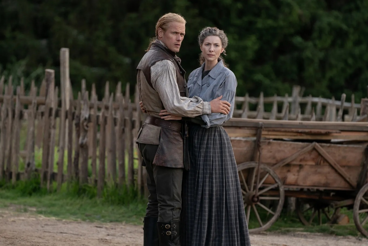 A white man with blond hair and a white woman with grey hair, both in simple period clothing, embrace as they stand on a dirt path with a wooden fence and cart in the background