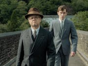 A young white in a light grey suit walks behind a middle aged white man in a dark grey suit and brown hat as they cross a stone bridge in the Welsh countryside
