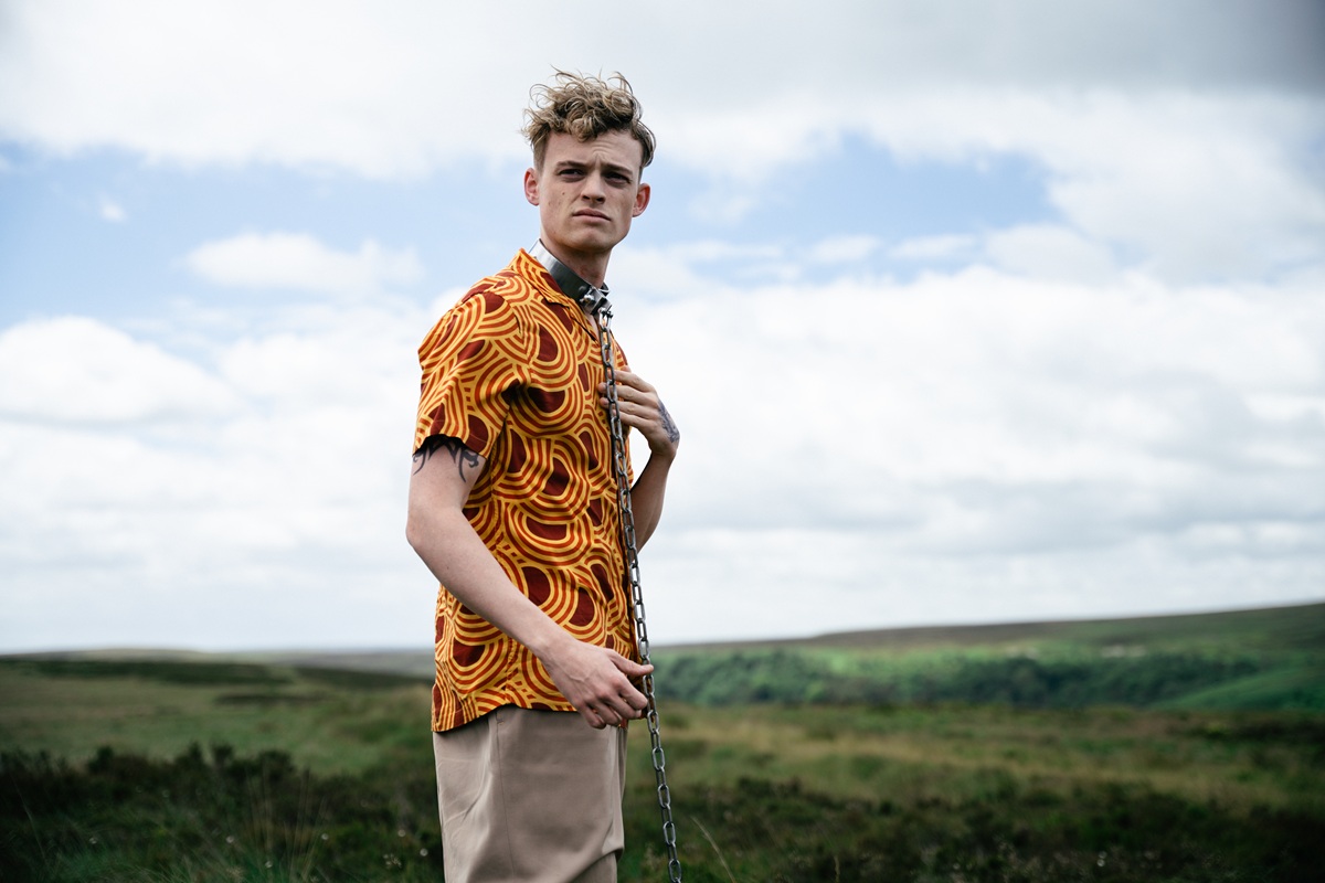 A young white man in a colourful shirt with a thick metal collar and chain around his neck, standing concerned in a bright field
