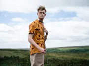 A young white man in a colourful shirt with a thick metal collar and chain around his neck, standing concerned in a bright field