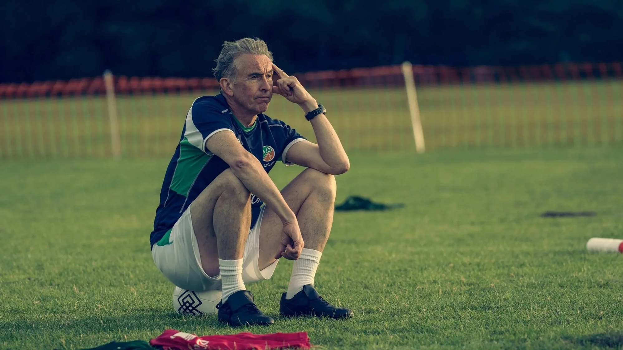 A middle aged white man in an Ireland Football kit sitting on a football pitch looking despondent