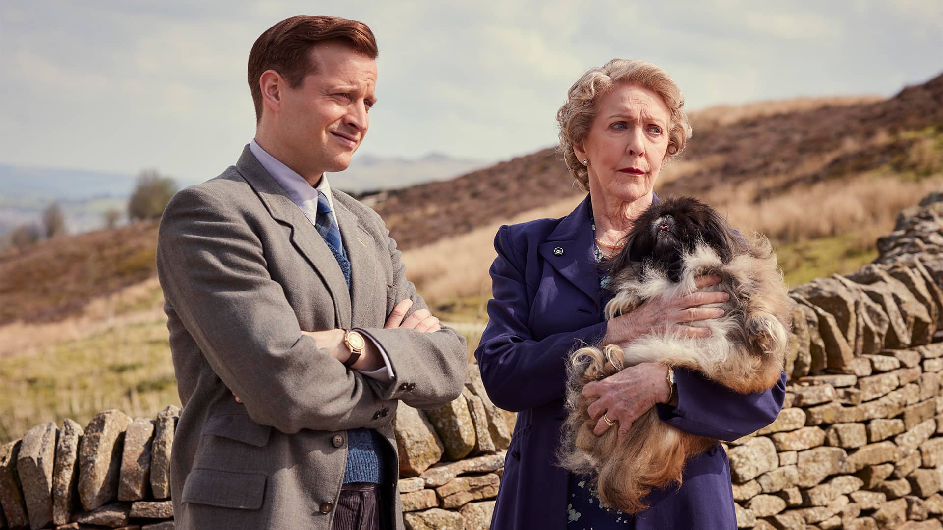 A white man and woman stand over the countryside. The woman holds a small dog.