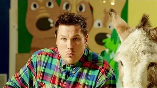 A white man in a colorful plaid shift has his lips pressed together as a donkey stands next to him.