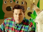 A white man in a colorful plaid shift has his lips pressed together as a donkey stands next to him.