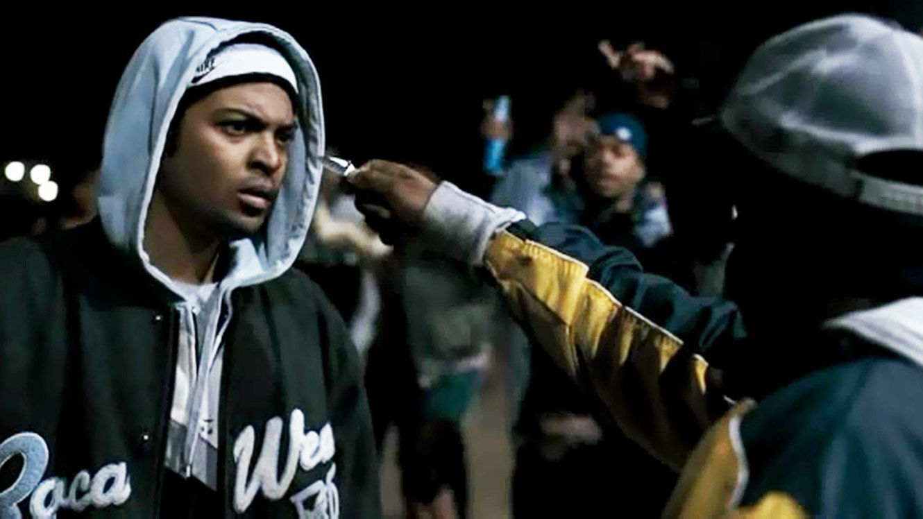 A black man stands in a hoodie with another man holding a knife towards his face.
