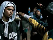 A black man stands in a hoodie with another man holding a knife towards his face.