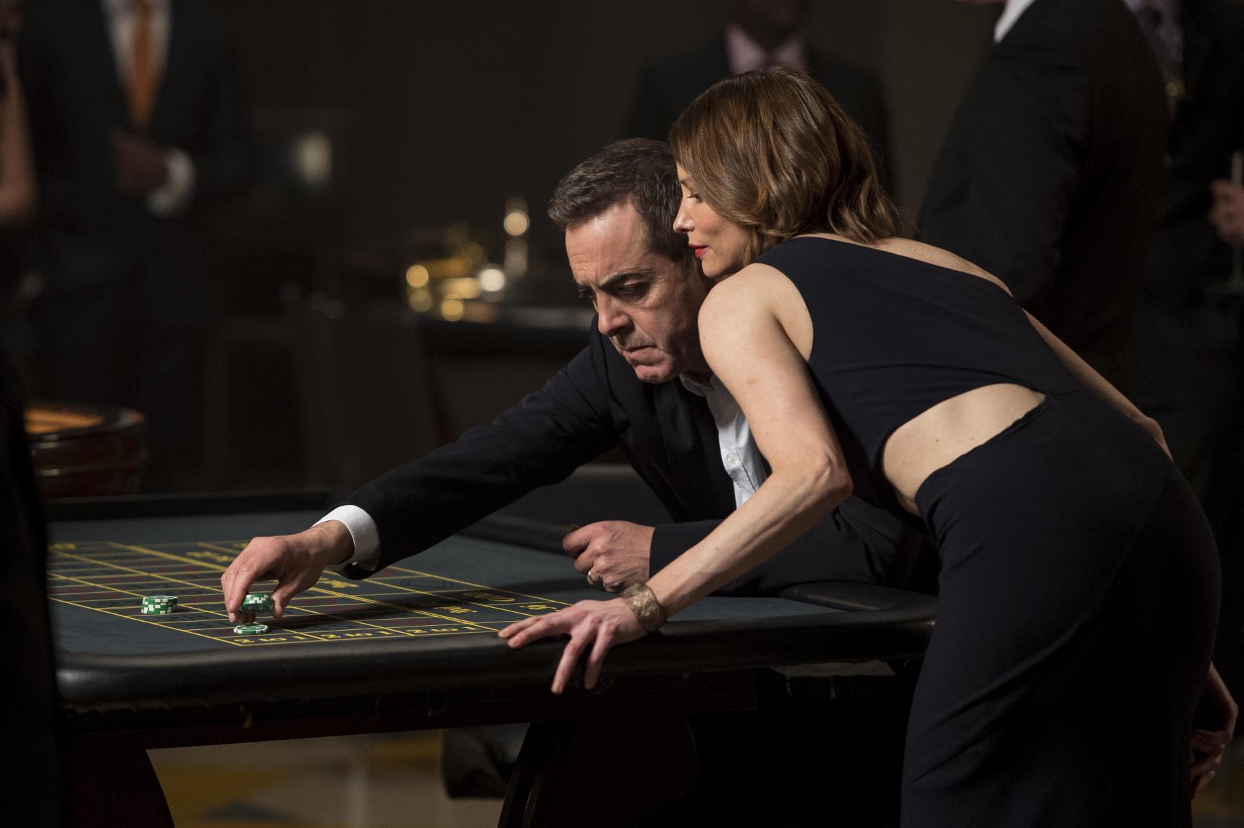 A white woman leans over a white man over a betting table. The man is placing green chips.