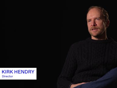 A white middle aged man in a black sweater and blue jeans sits giving an interview, with graphic text 'Kirk Hendry - Director' on the images