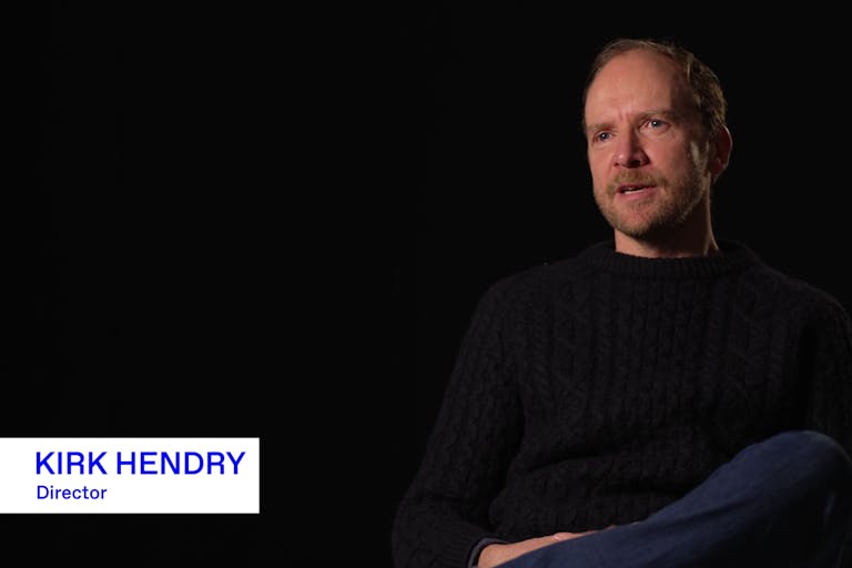 A white middle aged man in a black sweater and blue jeans sits giving an interview, with graphic text 'Kirk Hendry - Director' on the images