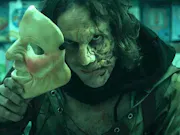 A man removes his mask revealing a slightly deformed face underneath.