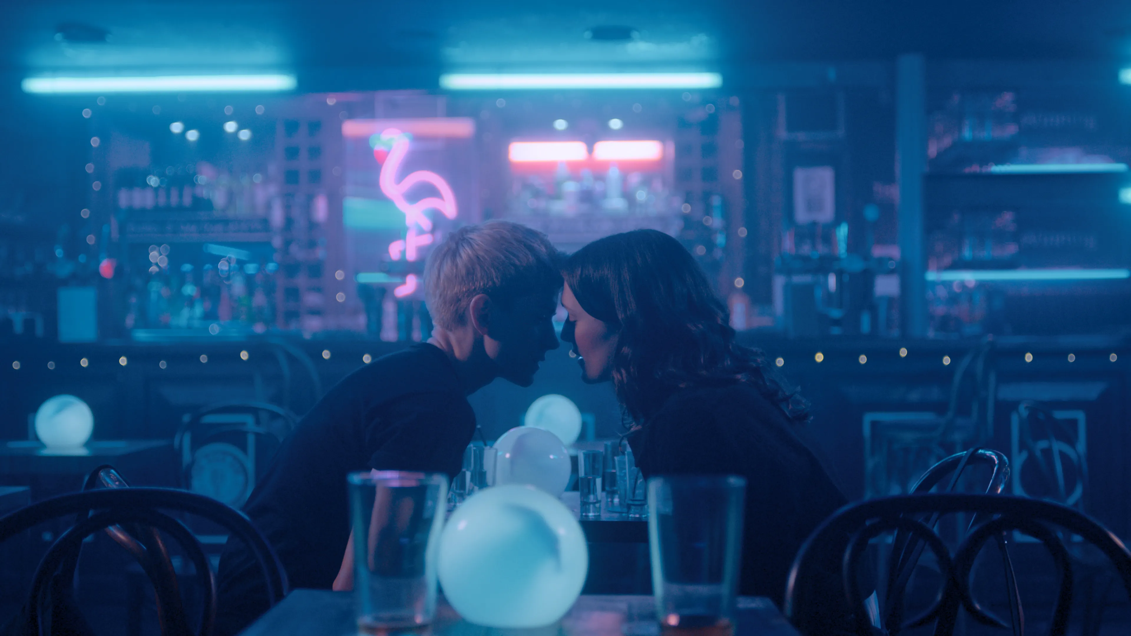 Two woman sitting in a blue-lit bar with their foreheads touching.