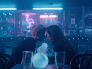 Two woman sitting in a blue-lit bar with their foreheads touching.