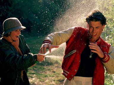 A woman gleefully sprays champagne on a shocked man wearing a sporting letterman jacket