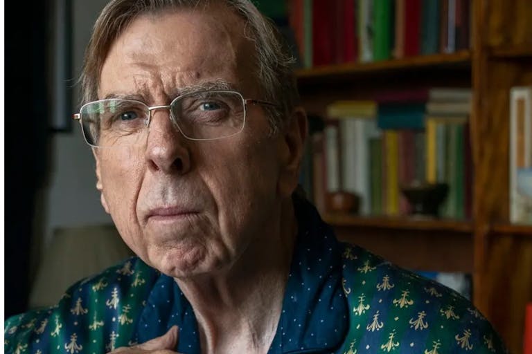 An older white man in glasses looking thoughtful, a bookcase behind him