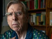 An older white man in glasses looking thoughtful, a bookcase behind him