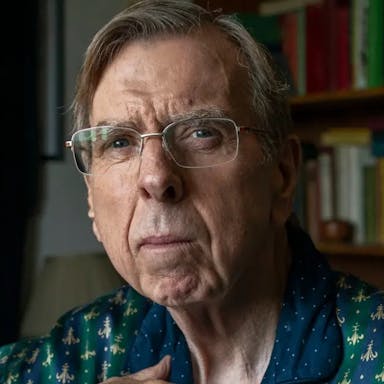 An older white man in glasses looking thoughtful, a bookcase behind him 