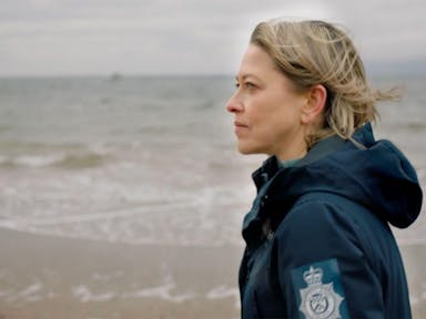 A white middle aged woman in a Police Detective coat standing on a windy beach shore looking thoughtful