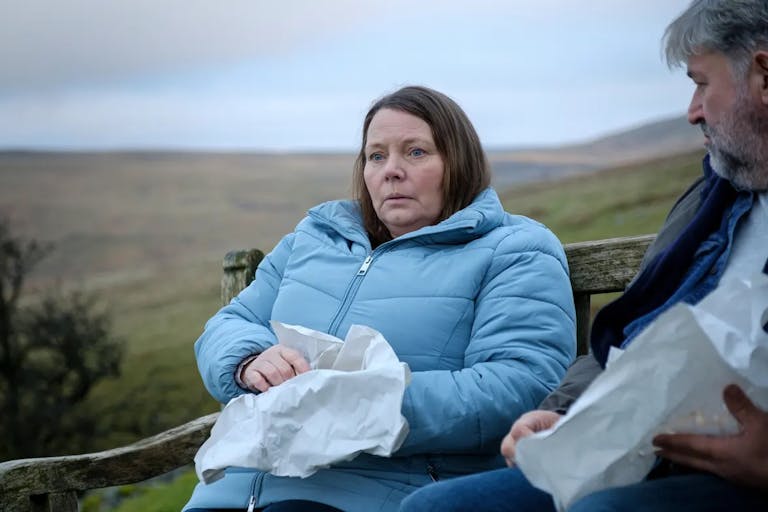 A middle aged white woman sits beside a middle aged white man on a bench in the countryside eating chips