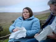 A middle aged white woman sits beside a middle aged white man on a bench in the countryside eating chips