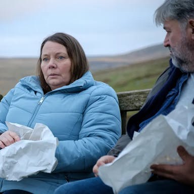 A middle aged white woman sits beside a middle aged white man on a bench in the countryside eating chips