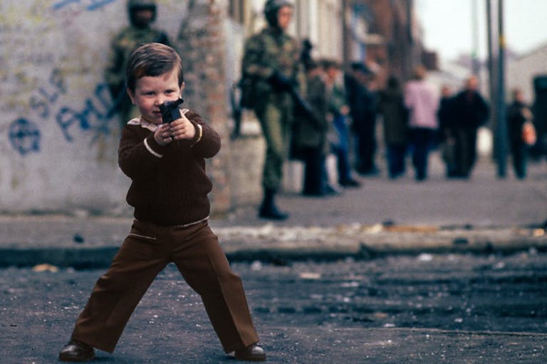 A young white boy wearing brown clothes in Northern Ireland playing with a fake gun while two fully armed up British soldiers walk in the background