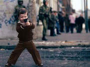 A young white boy wearing brown clothes in Northern Ireland playing with a fake gun while two fully armed up British soldiers walk in the background