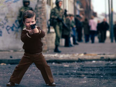 A young white boy wearing brown clothes in Northern Ireland playing with a fake gun while two fully armed up British soldiers walk in the background