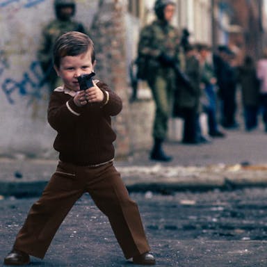 A young white boy wearing brown clothes in Northern Ireland playing with a fake gun while two fully armed up British soldiers walk in the background