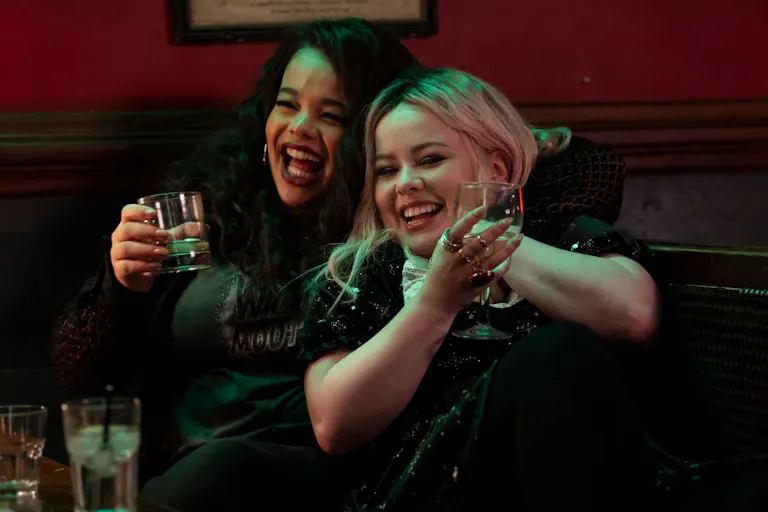 Two young women, one white one mixed race, holding drinks in a bar and laughing