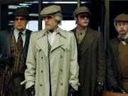 Four men stand in disguise wearing sunglasses, flat caps and fake beards.