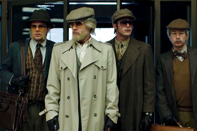 Four men stand in disguise wearing sunglasses, flat caps and fake beards.