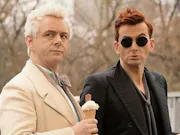 Two white men. One white haired and wearing a white suit, one red-haired and wearing a black suit and glasses. Both looking in the same direction
