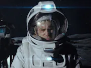 A man wearing a spacesuit on the moon, walking beside a large space drone