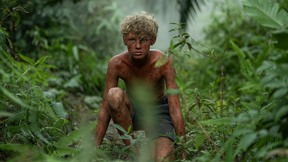 A boy covered in mud, crouching in a rainforest