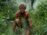 A boy covered in mud, crouching in a rainforest