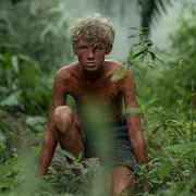 A boy covered in mud, crouching in a rainforest