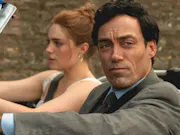 A suited man in the passenger seat of a convertible, while a red-haired woman drives