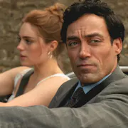 A suited man in the passenger seat of a convertible, while a red-haired woman drives