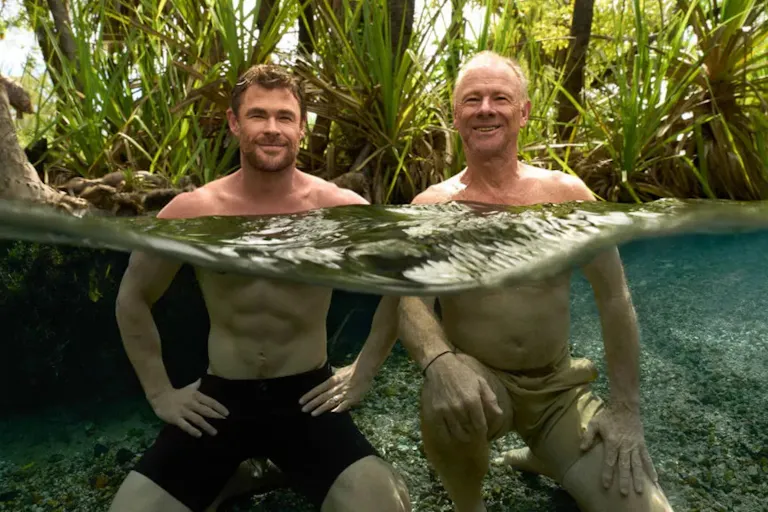 Two white men (one middle aged, one elderly) swimming in a body of water in a rainforest