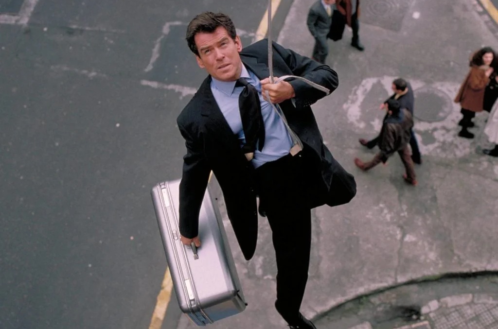 A white man in a suit hanging from a wire over a busy street, suitcase in hand