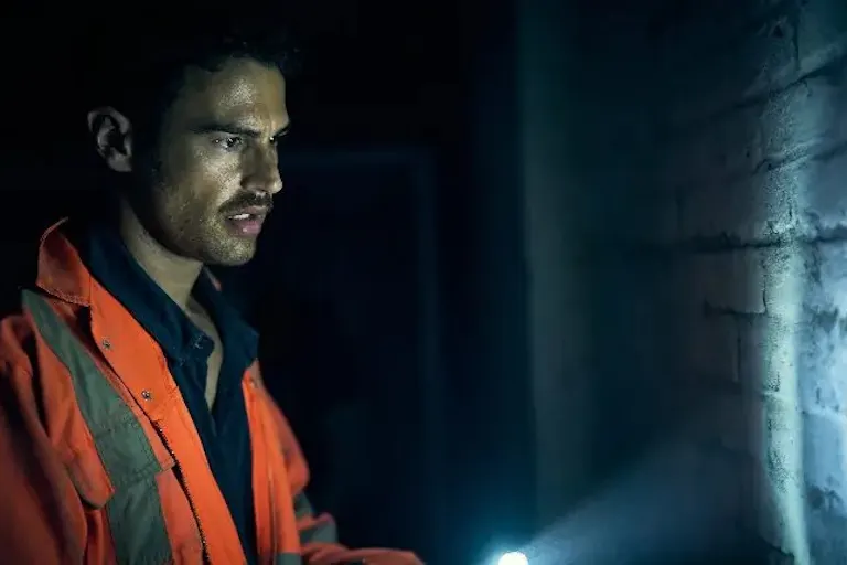 A white man wearing a high-vis jacket in a dark room, pointing a torchlight at a wall
