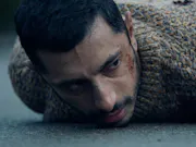 A close up of a south asian young man laying face-down on concrete, with blood on his cheek