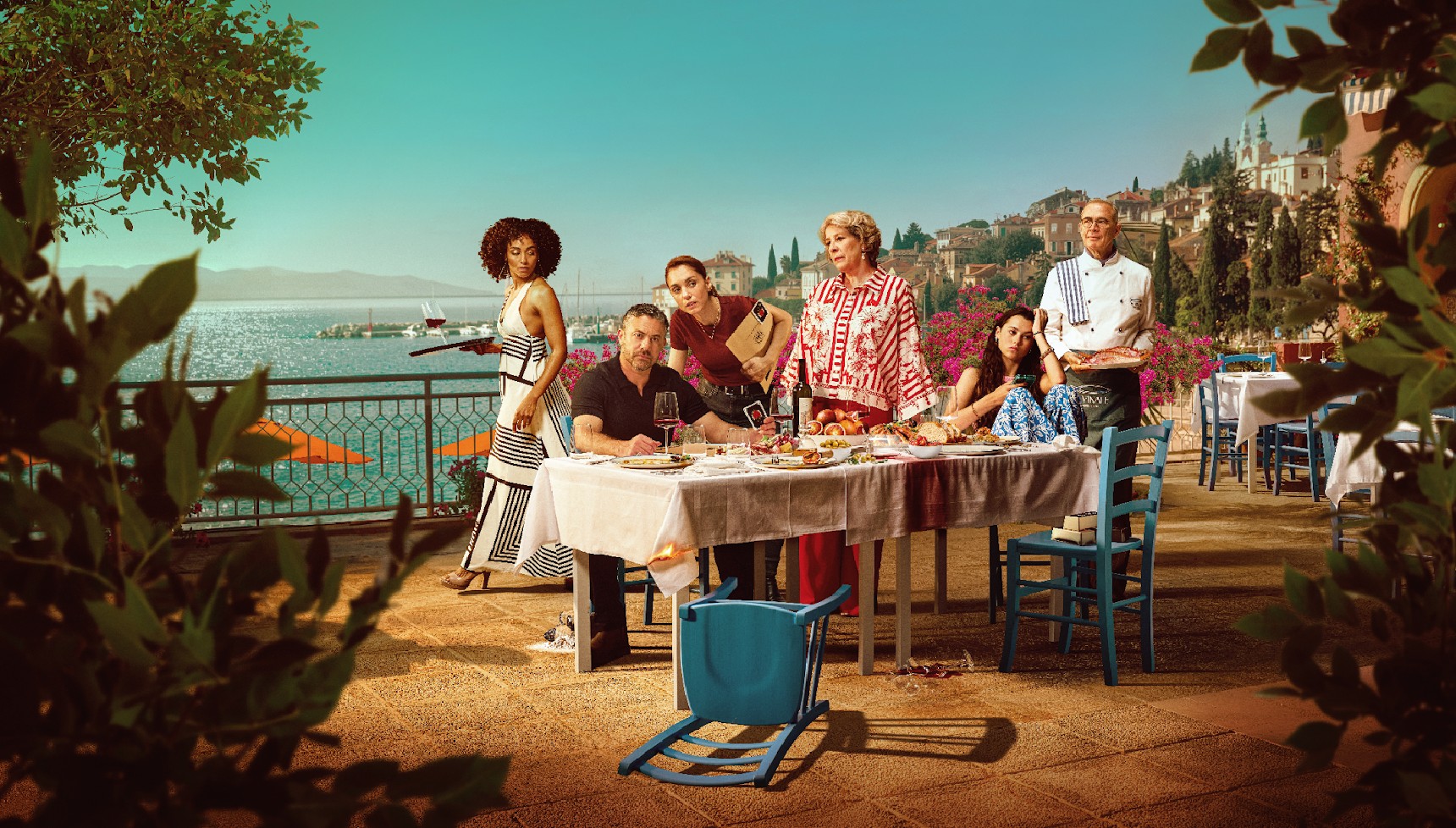 A group of people standing around a dinner table in summery clothing, within a sunny mediterranean setting